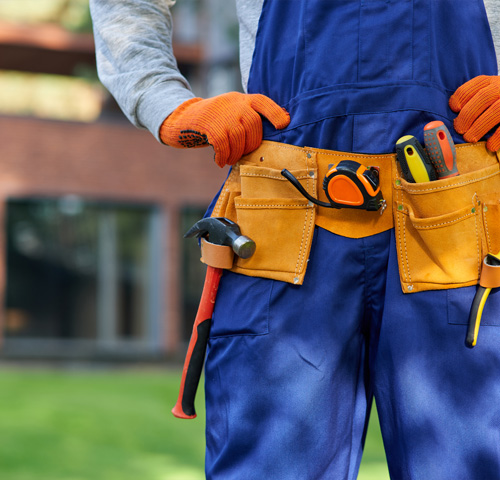 person wearing orange gloves and blue overalls with a tool belt containing various tools including a tape measure and hammer in a construction setting with green grass and a building in the background three essential tools for work